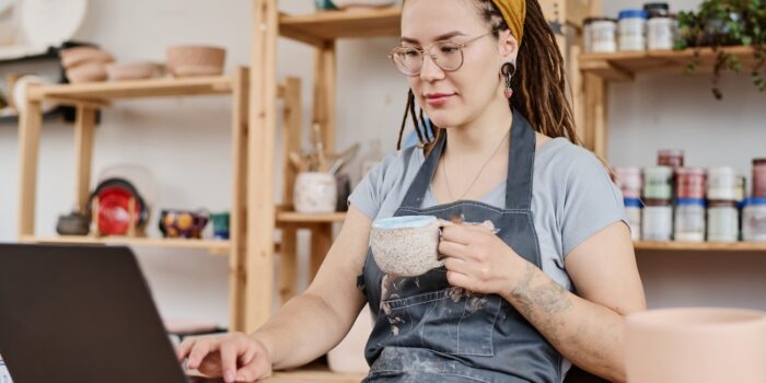 Young female freelancer or owner of small business sitting in front of laptop by workplace, having tea and searching for new ideas in the net